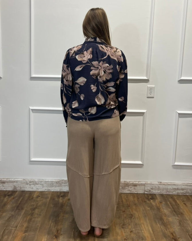 A woman viewed from behind wearing a navy blue floral long-sleeve blouse and beige wide-leg pleated pants, standing in front of a white paneled wall on a wood floor.