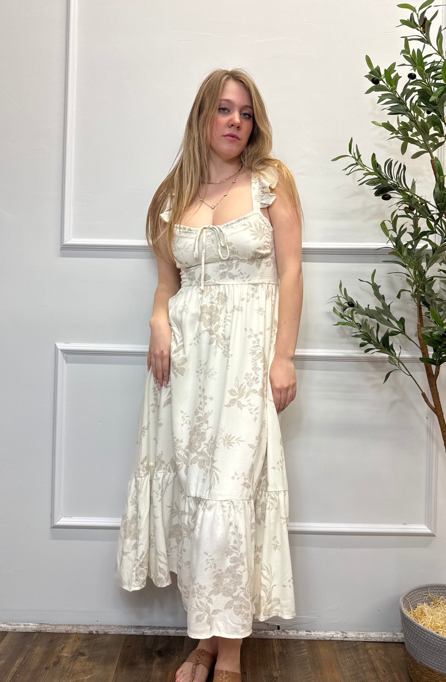 Woman wearing a white floral dress standing against a white wall with a plant to the side.
