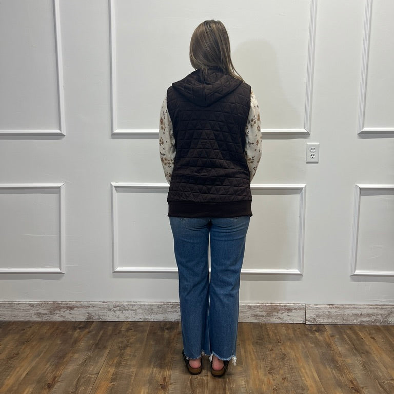 A woman stands with her back to the camera wearing a black quilted puffer vest over a patterned long-sleeve top and blue jeans, standing in a room with white paneled walls and wood floors.