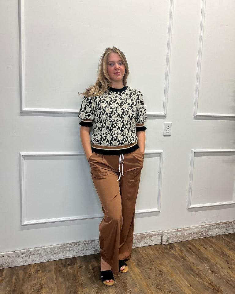 A woman stands indoors against a white paneled wall wearing a black-and-white patterned short-sleeve top, brown wide-leg pants, and black sandals.