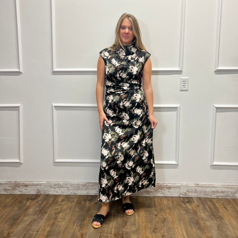 A woman modeling a sleeveless black midi dress with a white and gray floral pattern, standing against a white paneled wall on a wood floor.