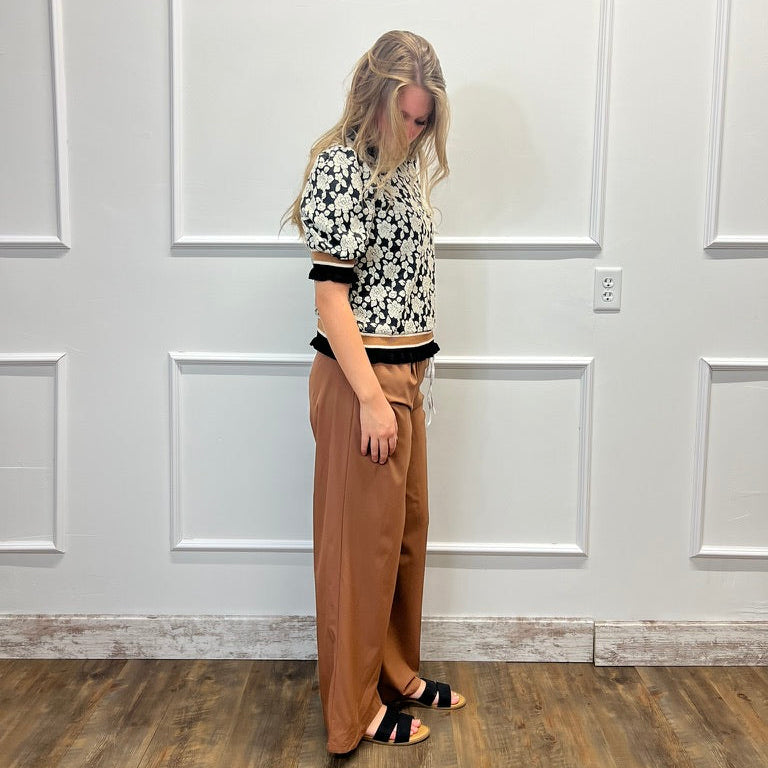 A woman stands in a room with white paneled walls and a wood floor, wearing a black-and-white patterned short-sleeve crop top, tan wide-leg pants, and black sandals.