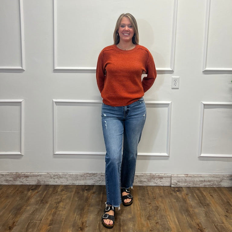 A woman wearing a rust-orange crewneck sweater and blue jeans stands against a white paneled wall on a wood floor.