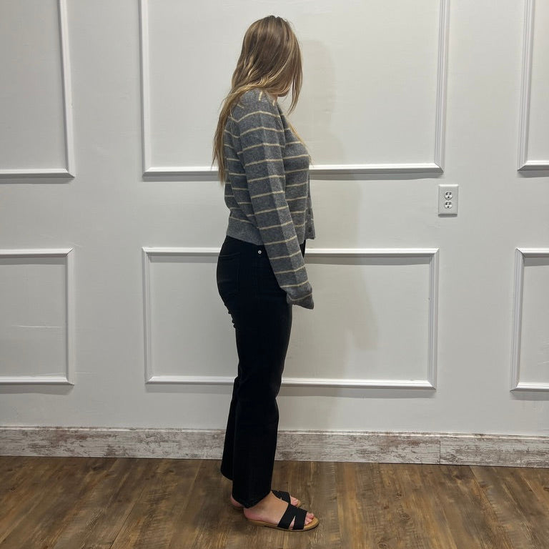A woman stands in profile wearing a gray and white striped long-sleeve knit top, black pants, and black slide sandals, standing on a wood floor against a white paneled wall.