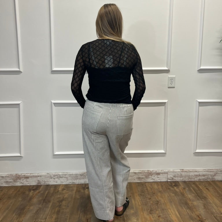 A woman viewed from behind wearing a black long-sleeve mesh top and light gray wide-leg jeans, standing on a wood floor against a white paneled wall.