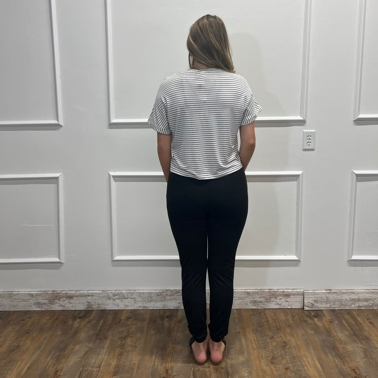 A woman viewed from behind wearing a white short-sleeve t-shirt with thin black horizontal stripes and black pants, standing on a wood floor against a white paneled wall.