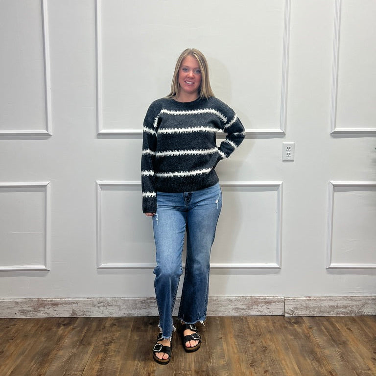 A woman stands in front of a white paneled wall wearing a black and white horizontally striped knit sweater, blue jeans, and black sandals.