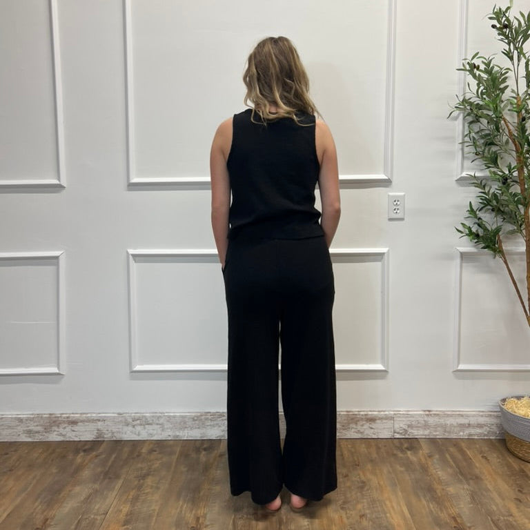 Person wearing a black sleeveless top and wide-leg pants standing in a room with white paneled walls and a wooden floor.