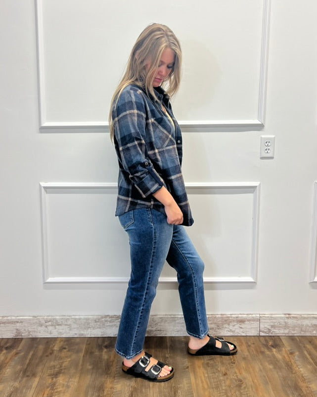 A woman stands in profile wearing a blue and black plaid flannel shirt, blue jeans, and black slide sandals against a white paneled wall with a wood floor.