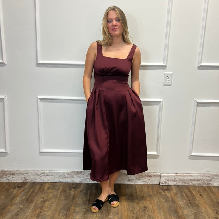 A woman modeling a sleeveless, scoop-neck midi dress in a deep burgundy satin fabric, standing against a white paneled wall with a wood floor.