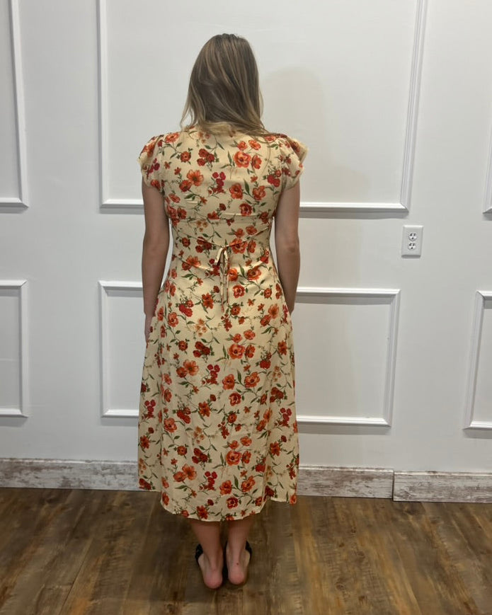 A woman viewed from behind wearing a knee-length cream dress with an orange and red floral pattern, standing in front of a white paneled wall on a wood floor.