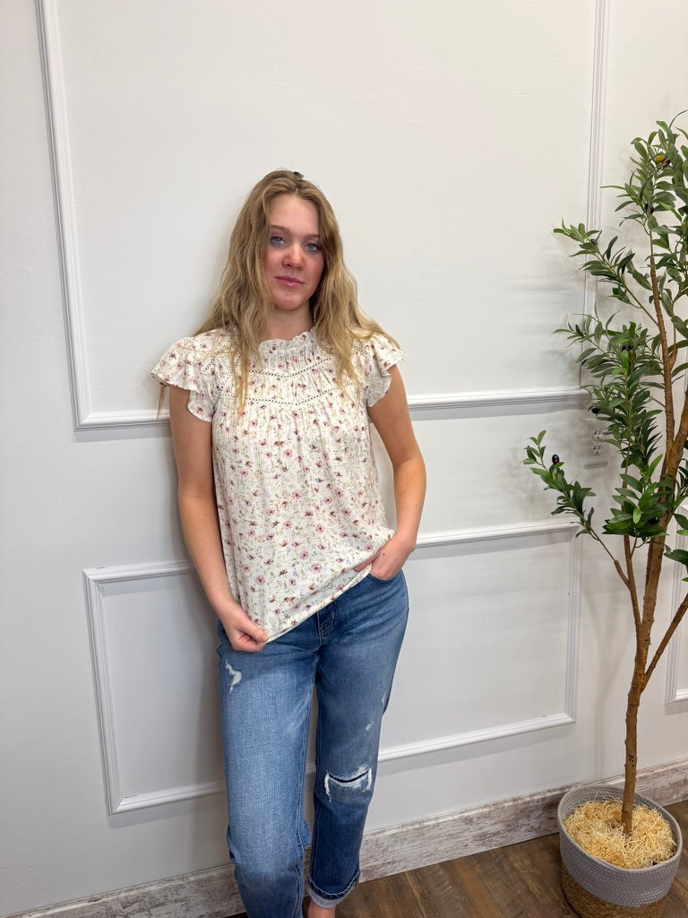 Woman wearing a floral blouse and jeans standing against a white paneled wall with a plant.