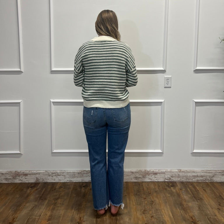 A woman viewed from behind wearing a green and white striped knit sweater and blue jeans, standing in front of a white paneled wall on a wood floor.