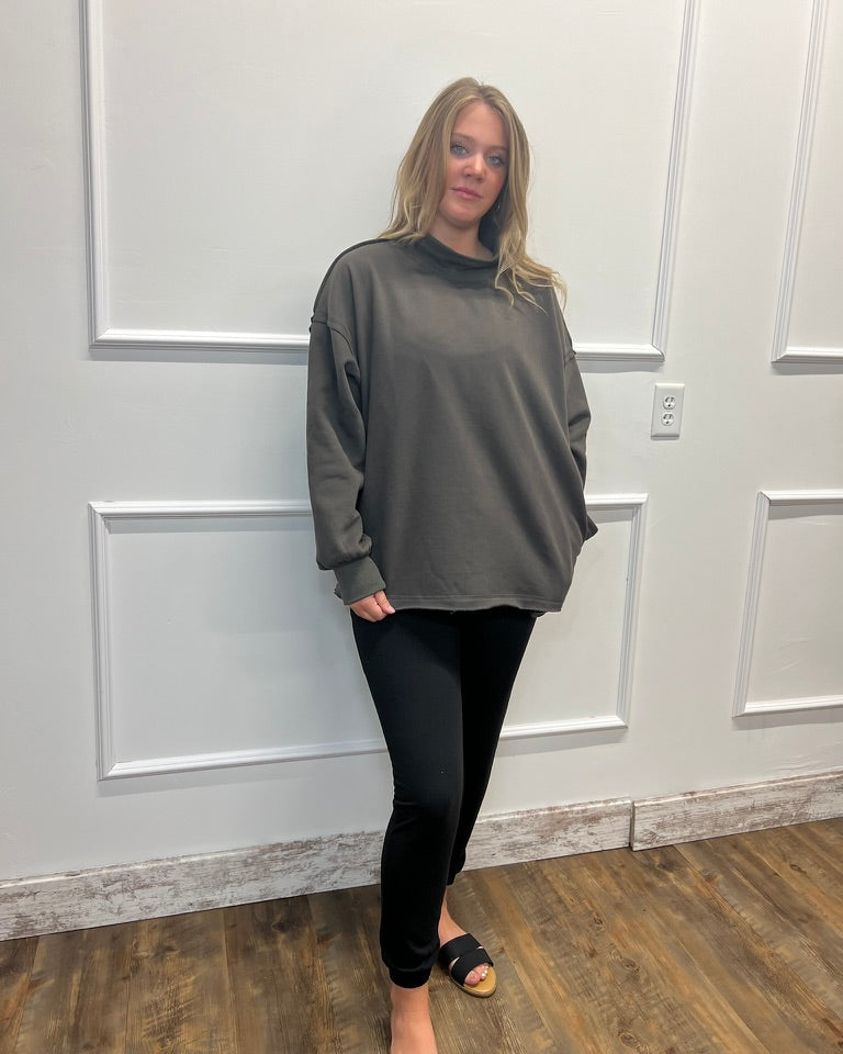 A woman modeling an oversized dark gray mock neck sweatshirt with black leggings and black slide sandals, standing in front of a white paneled wall.