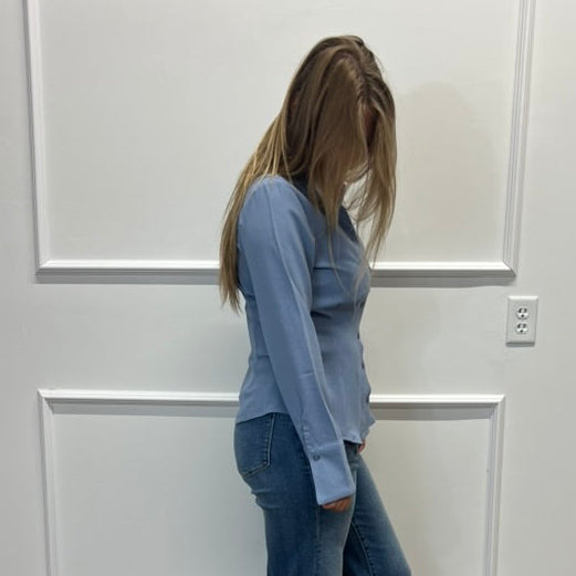A woman stands in profile wearing a light blue long-sleeve top and blue flared jeans, standing on a wood-look floor against a white paneled wall.
