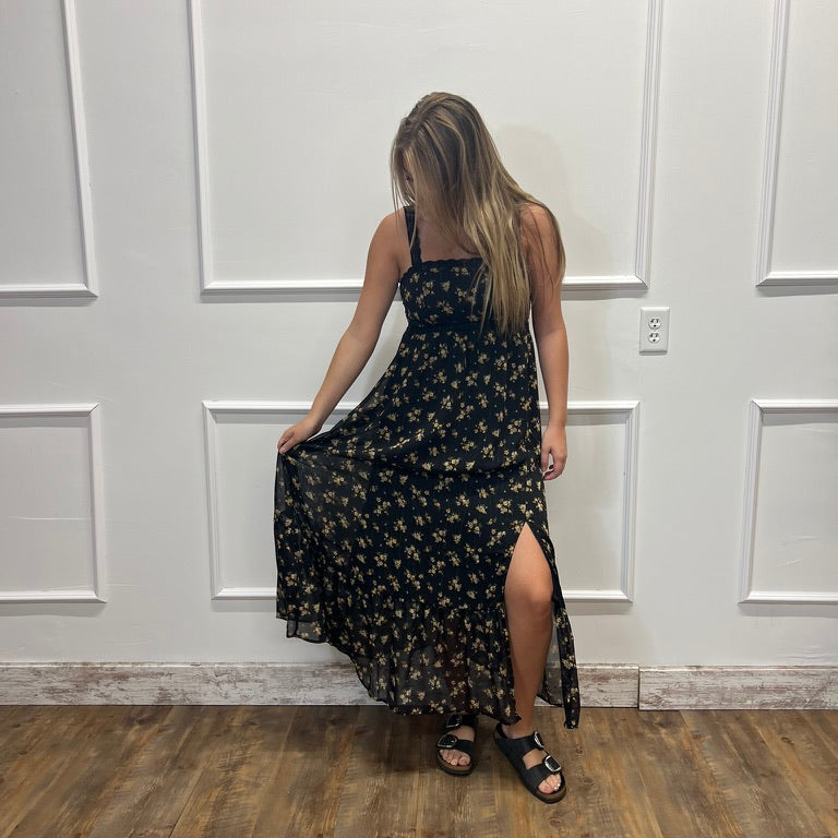 A woman wearing a black maxi dress with a small yellow and white floral print, standing with her back to the camera in front of a white paneled wall on a wood floor.