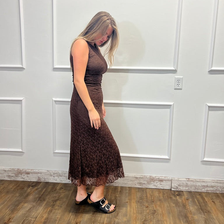 A woman modeling a sleeveless, floor-length brown lace dress in a room with white paneled walls and a wood floor.