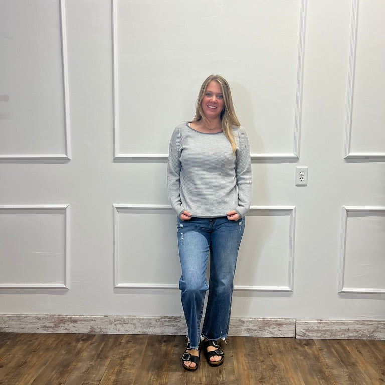 A woman stands in a room with white paneled walls and a wood floor, wearing a light gray long-sleeve knit sweater, blue jeans, and black sandals.