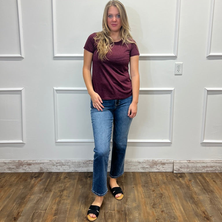 A woman stands in a room with white paneled walls and a wood floor, wearing a short-sleeve maroon t-shirt, blue jeans, and black slide sandals.
