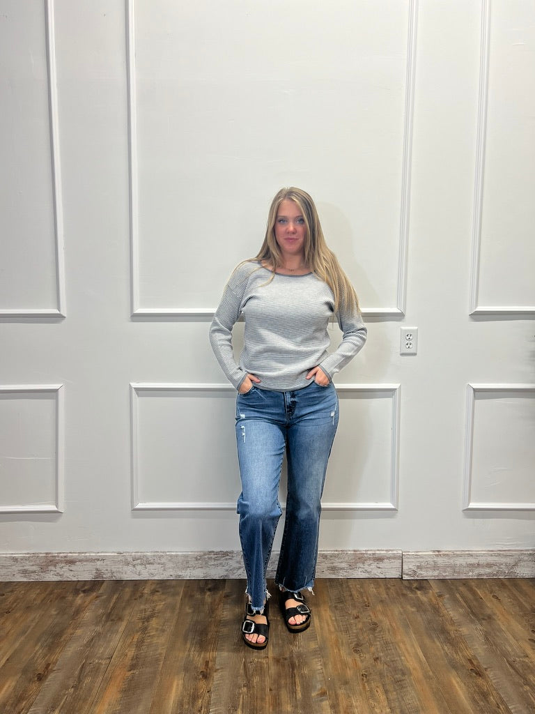 A woman stands indoors against a white paneled wall wearing a light gray long-sleeve top, blue jeans, and black sandals.