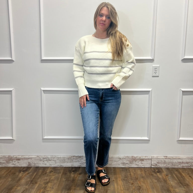 A woman stands in a room with white paneled walls and a wood floor, wearing a cream ribbed knit sweater, blue jeans, and black sandals.