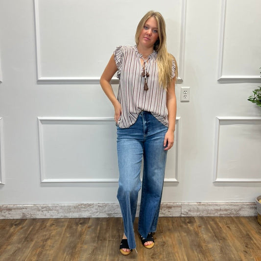 A woman modeling a sleeveless striped top with ruffled shoulders, paired with blue jeans and black sandals, standing in front of a white paneled wall on a wood floor.
