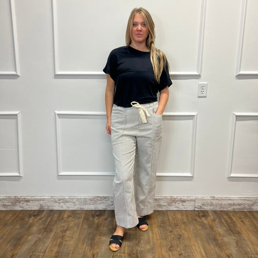 A woman stands in a room with white paneled walls and a wood floor, wearing a black short-sleeve t-shirt, light beige wide-leg pants with a cream drawstring, and black slide sandals.
