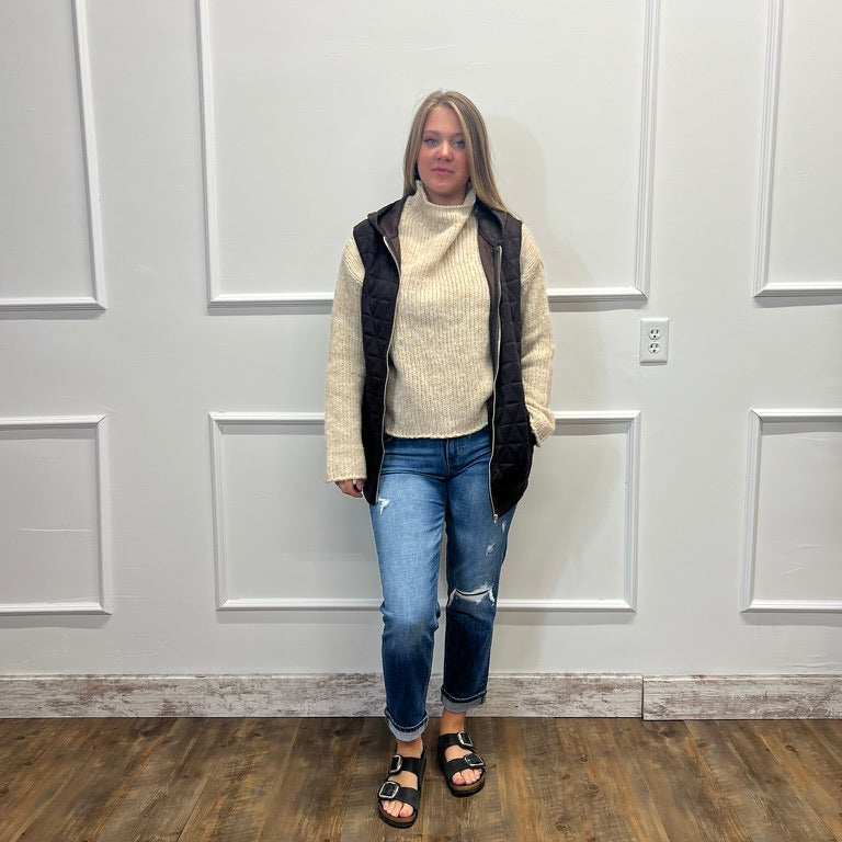 A woman stands in a room with white paneled walls and a wood floor, wearing a cream ribbed turtleneck sweater under a black quilted sleeveless vest, blue ripped jeans, and black slide sandals.