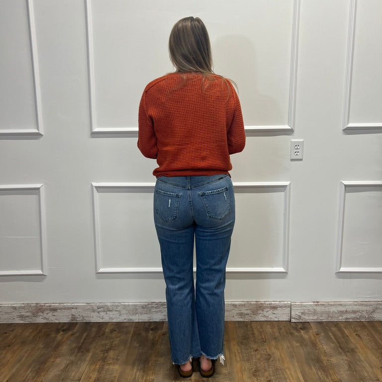 A woman viewed from behind wearing a rust-orange textured knit sweater and blue jeans, standing on a wood floor against a white paneled wall.