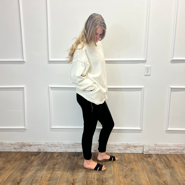 A woman stands in profile wearing a cream-colored oversized sweatshirt, black skinny pants, and black slide sandals, set against a white paneled wall and a wood-look floor.