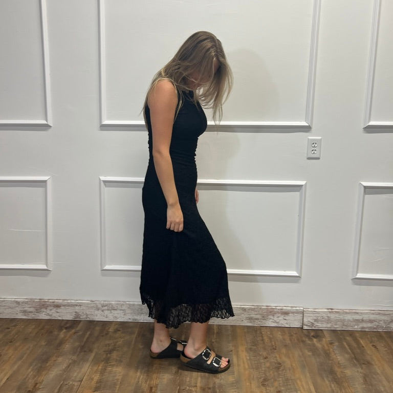 A woman wearing a sleeveless black midi dress with lace trim at the hem, standing in a room with white paneled walls and a wood floor.