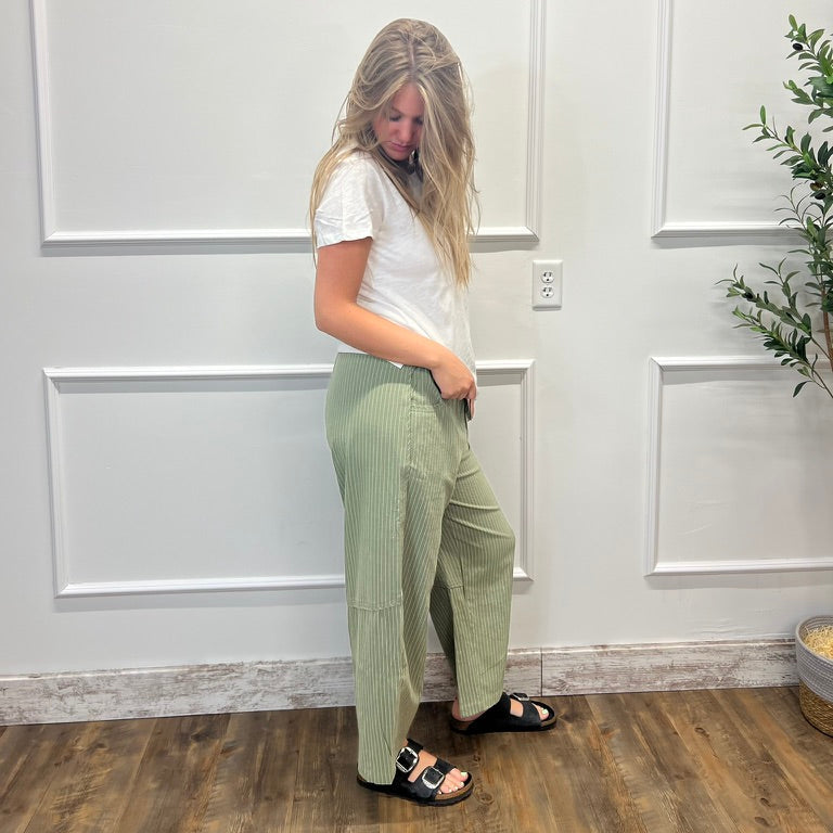 A woman with blonde hair stands in profile wearing light sage green pleated wide-leg trousers, a white t-shirt, and black slide sandals, posing against a white paneled wall with a wooden floor.