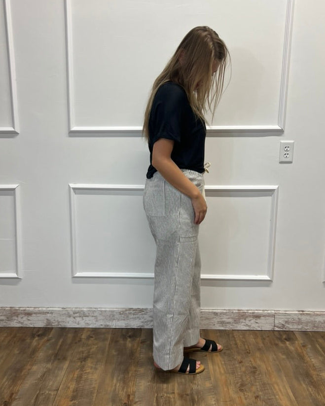 A woman stands in profile wearing a black short-sleeve top and light gray wide-leg pleated trousers, standing on a wood floor against a white paneled wall.