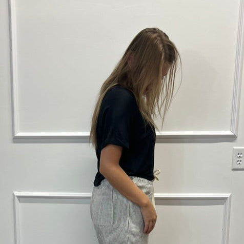 A woman stands in profile wearing a black short-sleeve top and light gray wide-leg pleated trousers, standing on a wood floor against a white paneled wall.