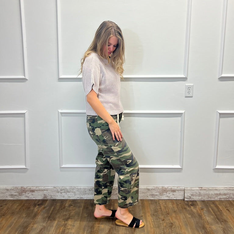 A woman stands in profile wearing camouflage pants, a light gray short-sleeve top, and black slide sandals against a white paneled wall with wood flooring.