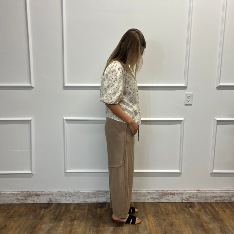 A woman stands in profile against a white paneled wall on a wood floor, wearing a cream floral short-sleeve blouse and beige wide-leg pants with black sandals.