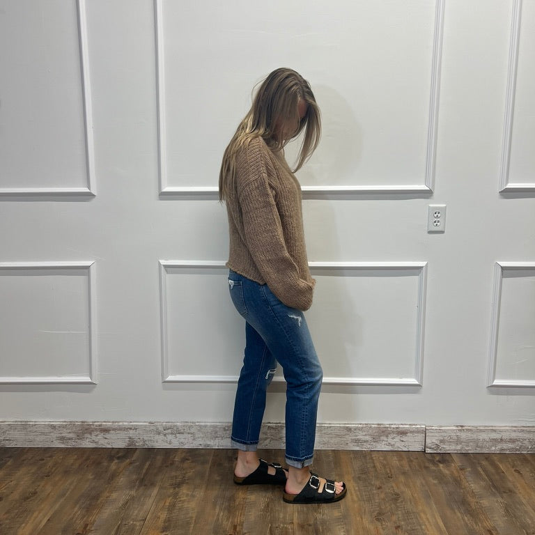 A woman stands in profile wearing a brown knit sweater, blue jeans, and black slide sandals, standing against a white paneled wall on a wood-look floor.