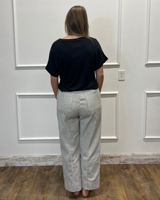 A woman stands with her back to the camera wearing a black short-sleeve top and light gray wide-leg trousers in a room with white paneled walls and a wooden floor.