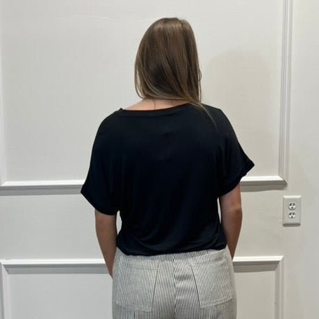 A woman stands with her back to the camera wearing a black short-sleeve top and light gray wide-leg trousers in a room with white paneled walls and a wooden floor.