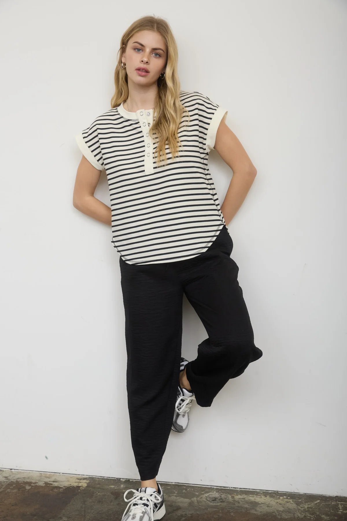 Woman wearing a black and white striped top and black pants against a white wall.