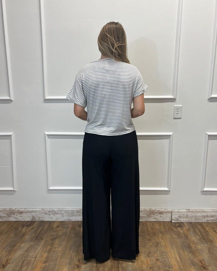 A woman stands with her back to the camera wearing a white t-shirt with thin black horizontal stripes and black wide-leg pants, standing in front of a white paneled wall on a wood floor.