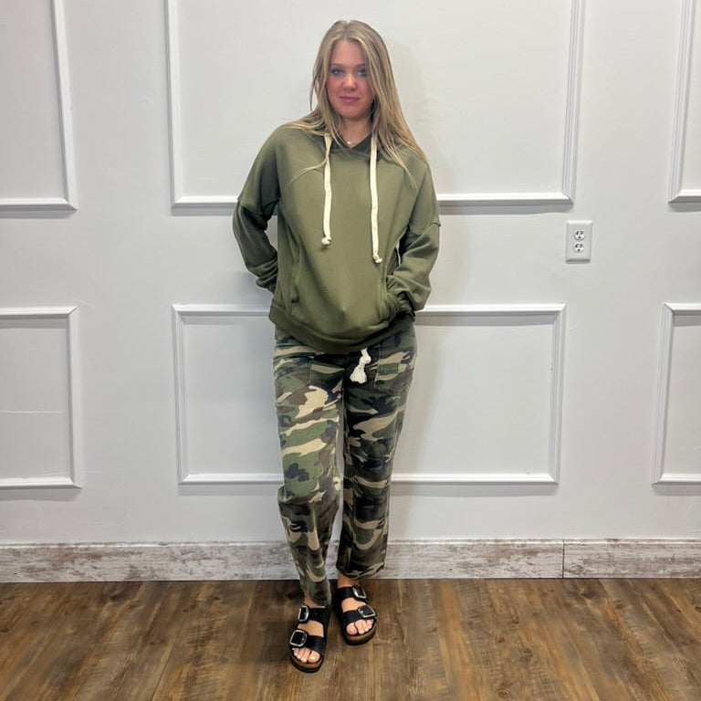 A woman stands indoors against a white paneled wall wearing an olive green hoodie with white drawstrings and matching camouflage jogger pants, paired with black slide sandals.