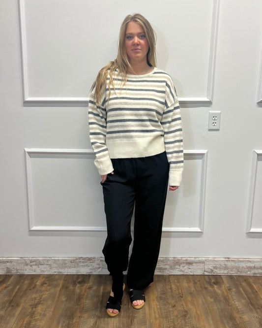 A woman stands in a room with white paneled walls and a wood floor, wearing a cream and gray striped knit sweater, black wide-leg pants, and black sandals.
