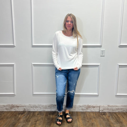 A woman stands against a white paneled wall wearing a white long-sleeve crewneck top, blue ripped jeans, and black slide sandals.