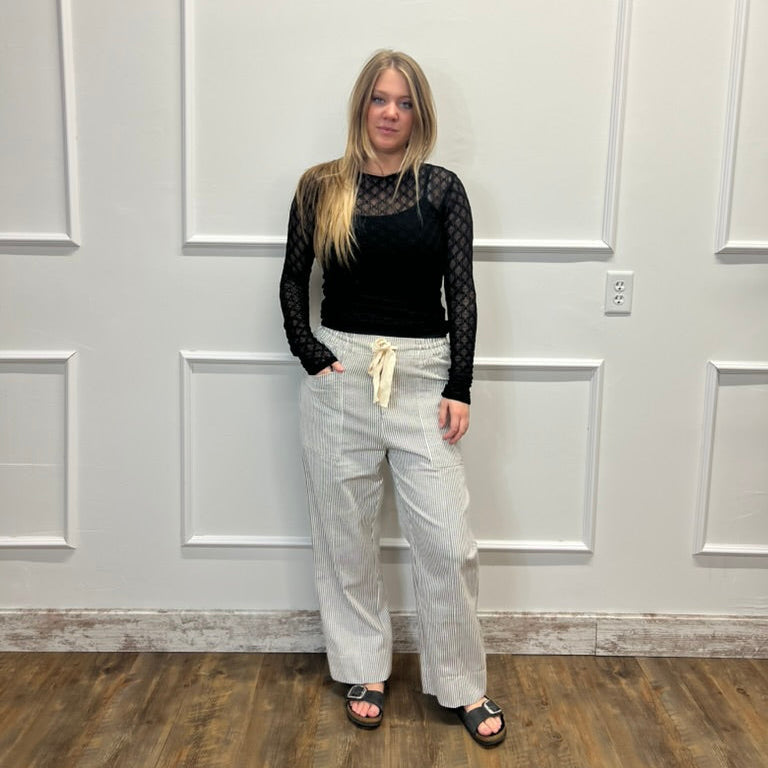 A woman stands in a room with white paneled walls and a wood floor, wearing a black long-sleeve top with sheer black mesh sleeves and light beige wide-leg pants.