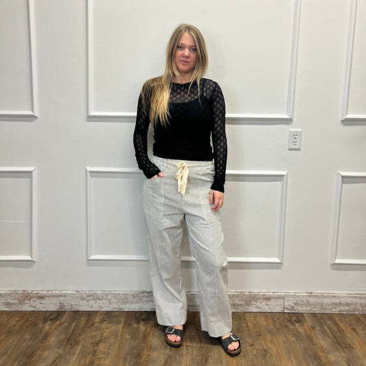 A woman stands in a room with white paneled walls and a wood floor, wearing a black long-sleeve top with sheer black mesh sleeves and light beige wide-leg pants.