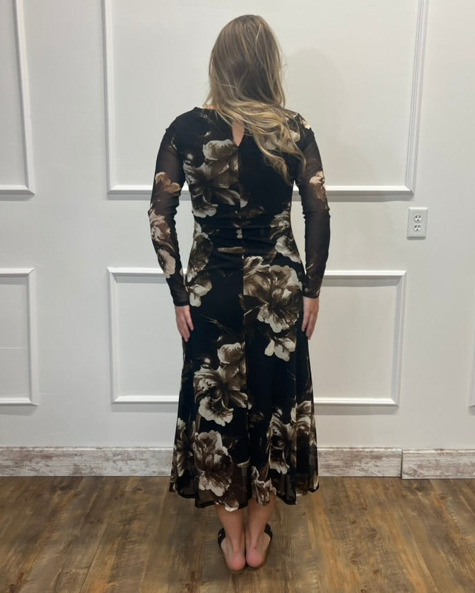 A woman stands with her back to the camera wearing a long-sleeve black midi dress with a brown and beige floral pattern, set against a white paneled wall and wood floor.