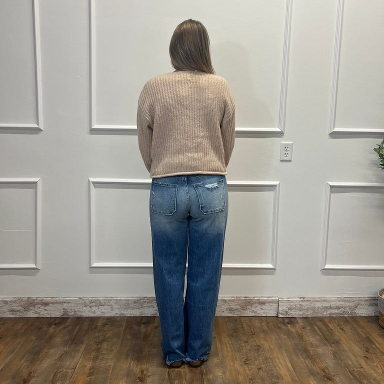 A person standing with their back to the camera, wearing a beige ribbed knit sweater and blue wide-leg jeans, positioned in front of a white paneled wall on a wooden floor.