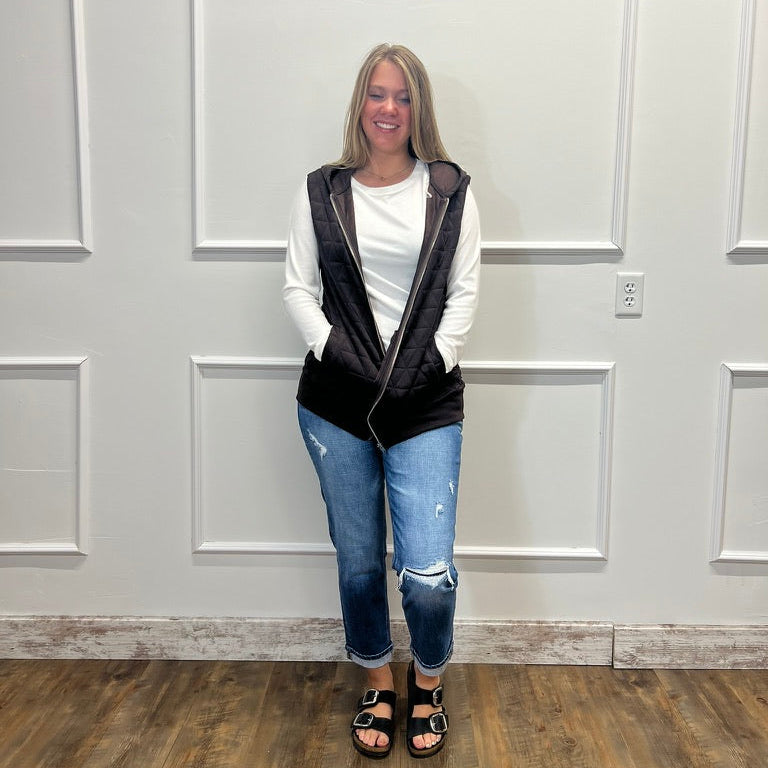 A woman stands indoors against a white paneled wall wearing a black zip-up vest over a white long-sleeve top, blue ripped jeans, and black sandals.
