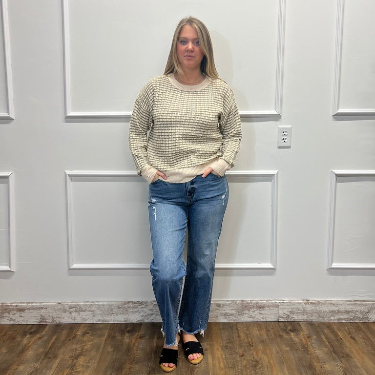 A woman stands in front of a white paneled wall wearing a beige and white textured knit sweater, blue jeans, and black sandals.